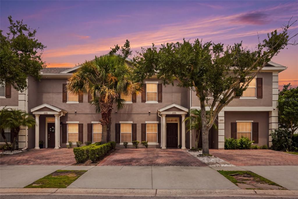 A multi-unit townhouse exterior with a brick driveway, palm trees, and a sunset sky — 2402 CARAVELLE CIRCLE, KISSIMMEE, FL