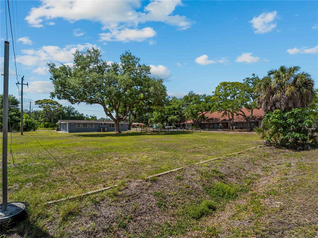 A large, grassy vacant lot features mature trees and nearby buildings under a blue sky — DAWN DRIVE, VENICE, FL