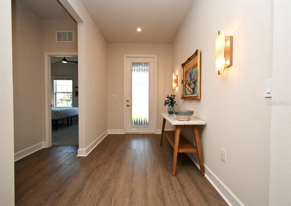 An entryway features wood-look flooring, a console table, wall sconces, and a view into a bedroom — 6650 PEBBLEBROOKE WAY, ZEPHYRHILLS, FL