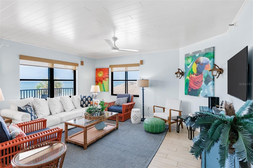 A bright living room features white plank ceilings, rattan furniture, and a view of the ocean — 17334 GULF BOULEVARD UNIT 203, NORTH REDINGTON BEACH, FL