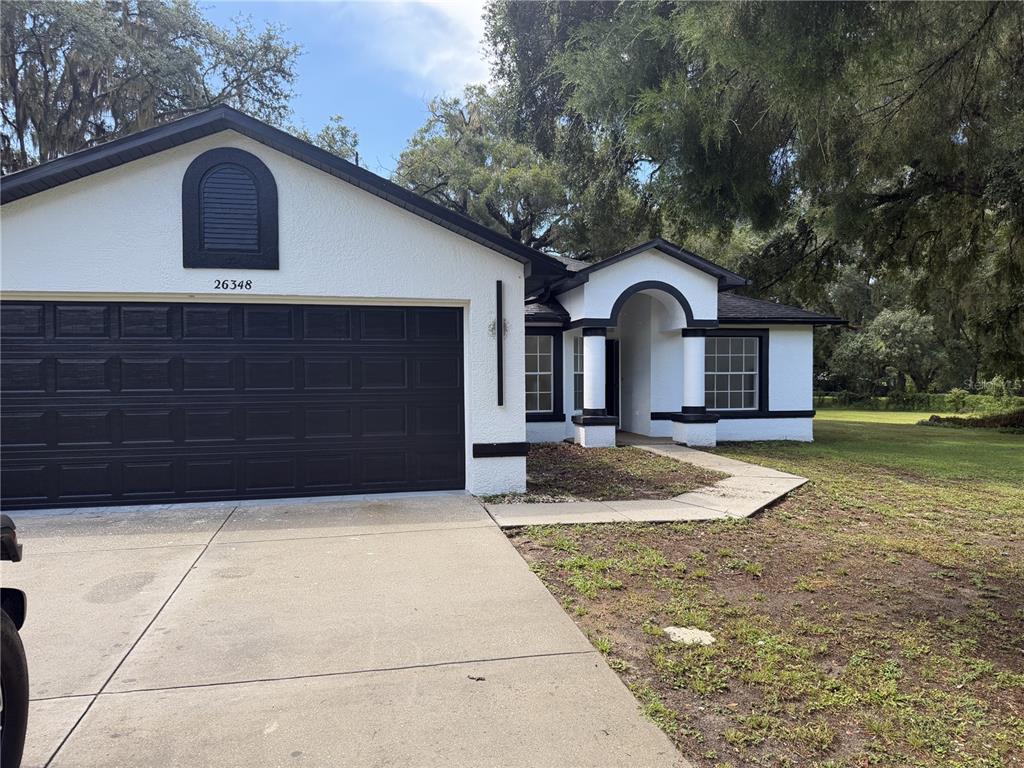 A white stucco house with a black garage door and black trim under large trees — 26348 CROOKED TREE LANE, BROOKSVILLE, FL