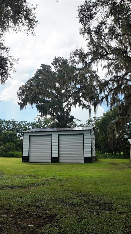 A detached metal garage with two roll-up doors stands in a grassy yard under trees — 26348 CROOKED TREE LANE, BROOKSVILLE, FL
