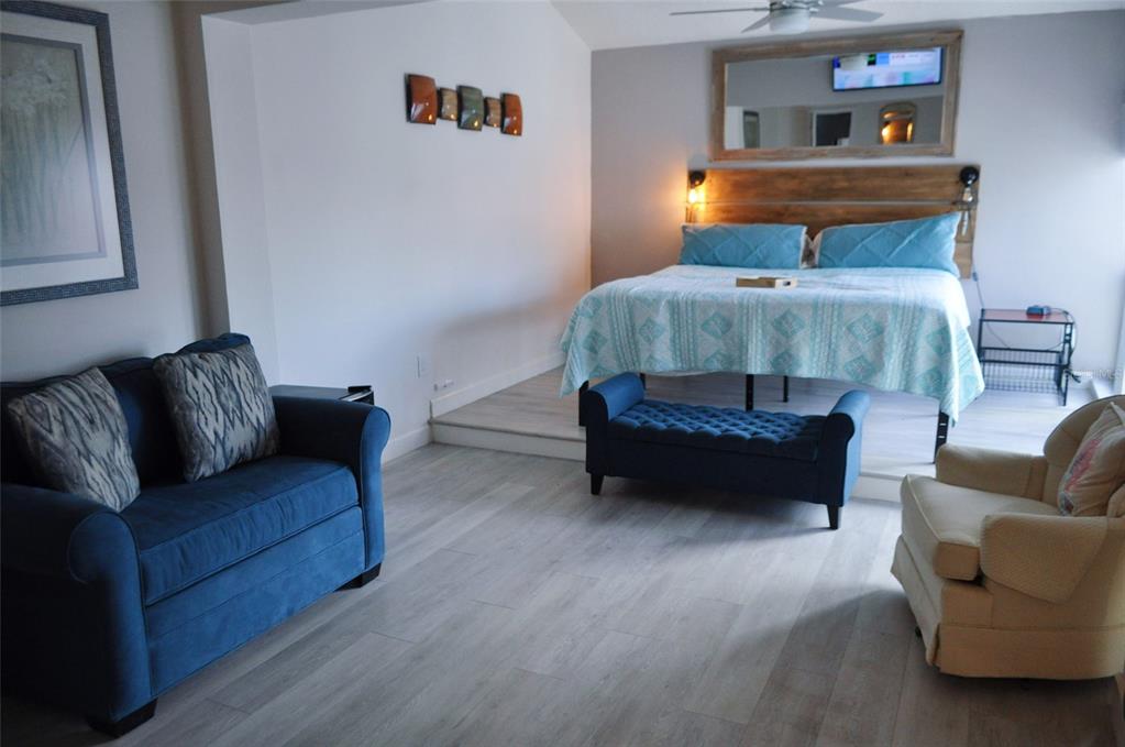 A bedroom with a raised bed area, blue sofa, armchair, and light wood-look flooring — 317 REX PLACE UNIT G, MADEIRA BEACH, FL
