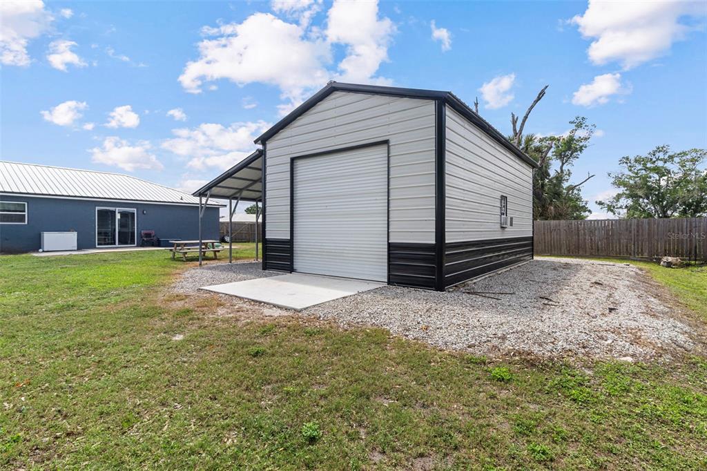 A detached metal garage with a side carport sits on a gravel lot behind a house — 1620 SE HAVEN DRIVE, ARCADIA, FL