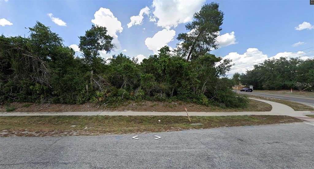 A vacant, heavily wooded corner lot sits behind a sidewalk along a paved road — 1850 PROVIDENCE BOULEVARD, DELTONA, FL