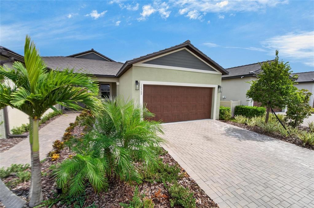 A single-story house features a paved driveway, a brown garage door, and tropical landscaping — 12569 LILY QUARTZ LOOP, PARRISH, FL