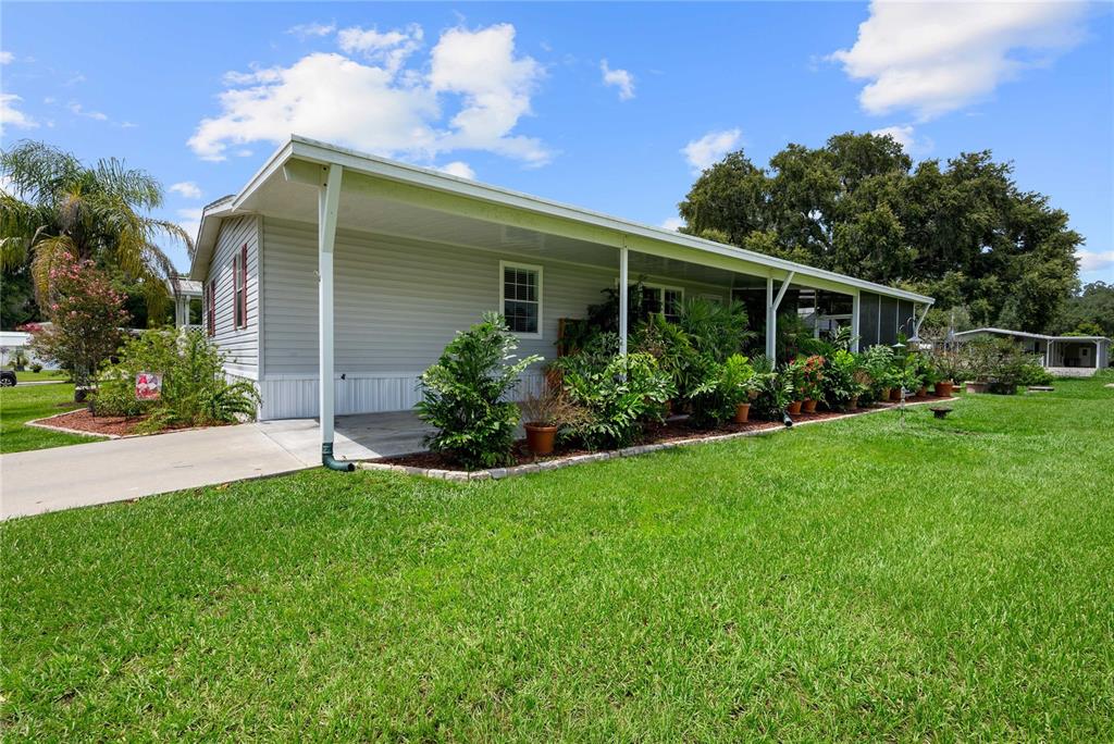 A light gray manufactured home features a long covered carport and a landscaped front yard — 38417 MONET DRIVE, ZEPHYRHILLS, FL