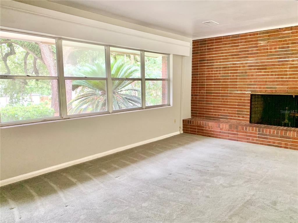 A living room features a brick fireplace, gray carpeting, and large windows overlooking trees — 2238 NW 16TH AVENUE, GAINESVILLE, FL