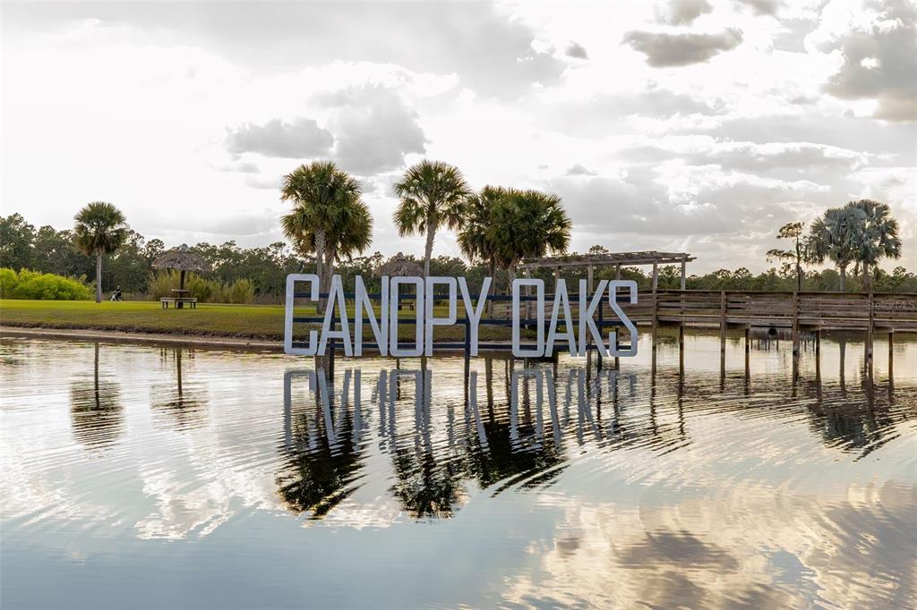 A sign reading Canopy Oaks stands in a lake in front of a wooden dock — 94 CEDAR CIRCLE E, LAKE WALES, FL
