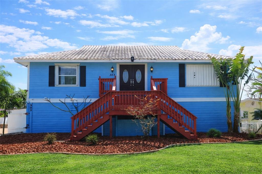 A bright blue elevated house features a central red wooden staircase leading to the entrance — 303 LAGOON DRIVE, PALM HARBOR, FL