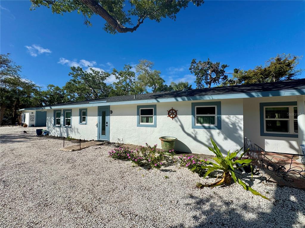A single-story white house with blue trim and a gravel yard under a blue sky — 7372 MELALEUCA WAY, SARASOTA, FL