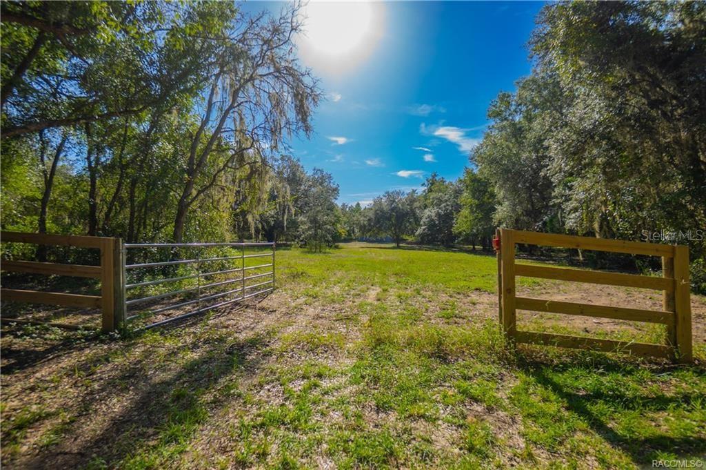 A metal gate opens into a grassy, tree-lined lot under a bright sun — 8929 E FLORAL PARK DRIVE, FLORAL CITY, FL
