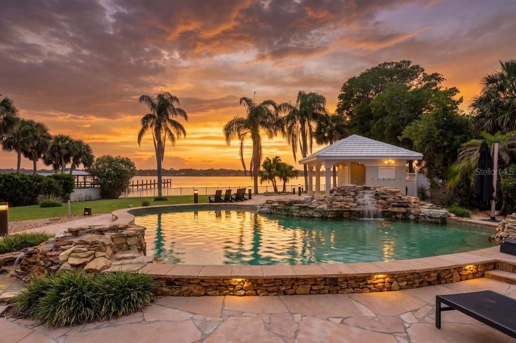 A swimming pool with a rock waterfall sits before a lake at sunset — 626 RIVERSIDE DRIVE, ORMOND BEACH, FL