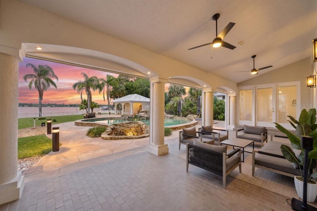 A covered patio features outdoor seating overlooking a swimming pool, waterfall, and waterfront sunset view — 626 RIVERSIDE DRIVE, ORMOND BEACH, FL