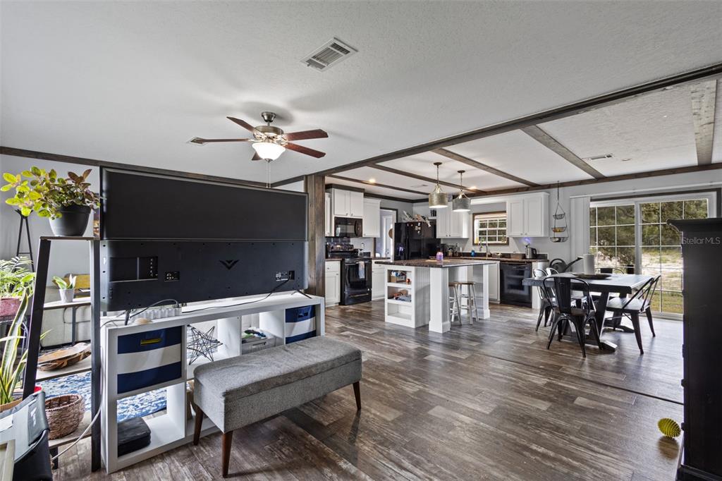 An open-concept kitchen and living area features wood-look flooring, exposed ceiling beams, and white cabinetry — 9490 NE 77TH PLACE, BRONSON, FL