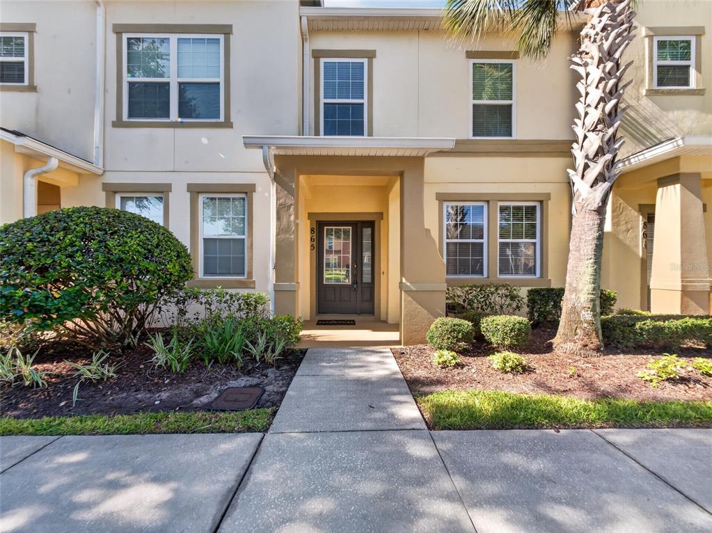 The exterior of a two-story townhouse features a tan facade, a covered entryway, and landscaping — 865 BELLES FERRY LOOP, SANFORD, FL