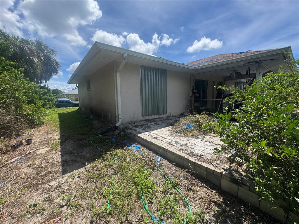 The exterior of a house features a concrete patio, metal storm shutters, and overgrown landscaping — 168 MILLPORT STREET NW, PORT CHARLOTTE, FL