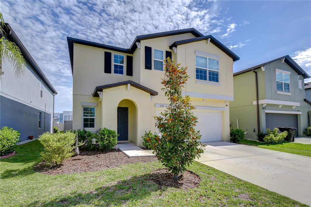 A two-story suburban house with a light yellow exterior, a driveway, and a small tree — 17917 CLOUDLESS BLISS DRIVE, LUTZ, FL