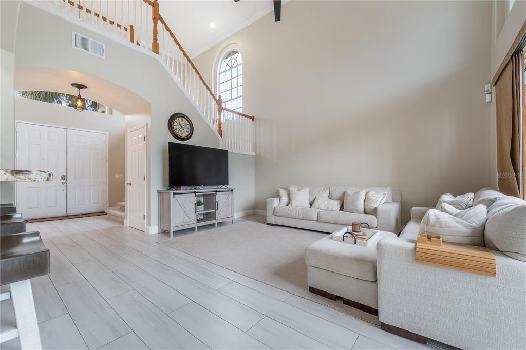 A bright living room features high ceilings, a staircase, light-colored tile flooring, and neutral furniture — 1524 THE OAKS BOULEVARD, KISSIMMEE, FL