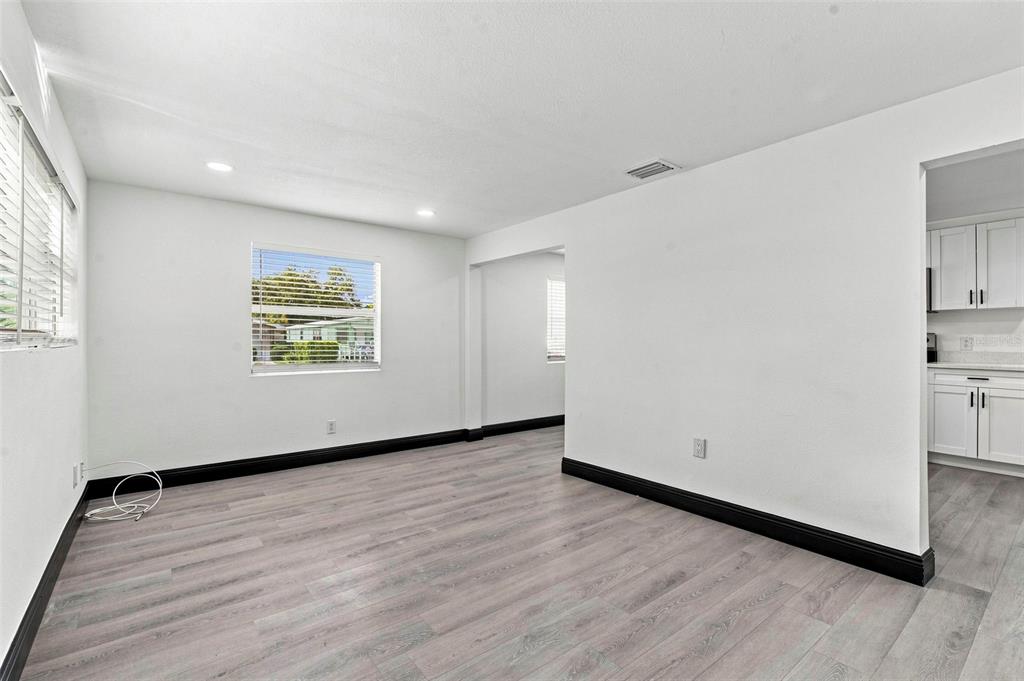 An empty room with light wood-look flooring, white walls, black baseboards, and a kitchen view — 3611 3RD AVENUE S, ST PETERSBURG, FL