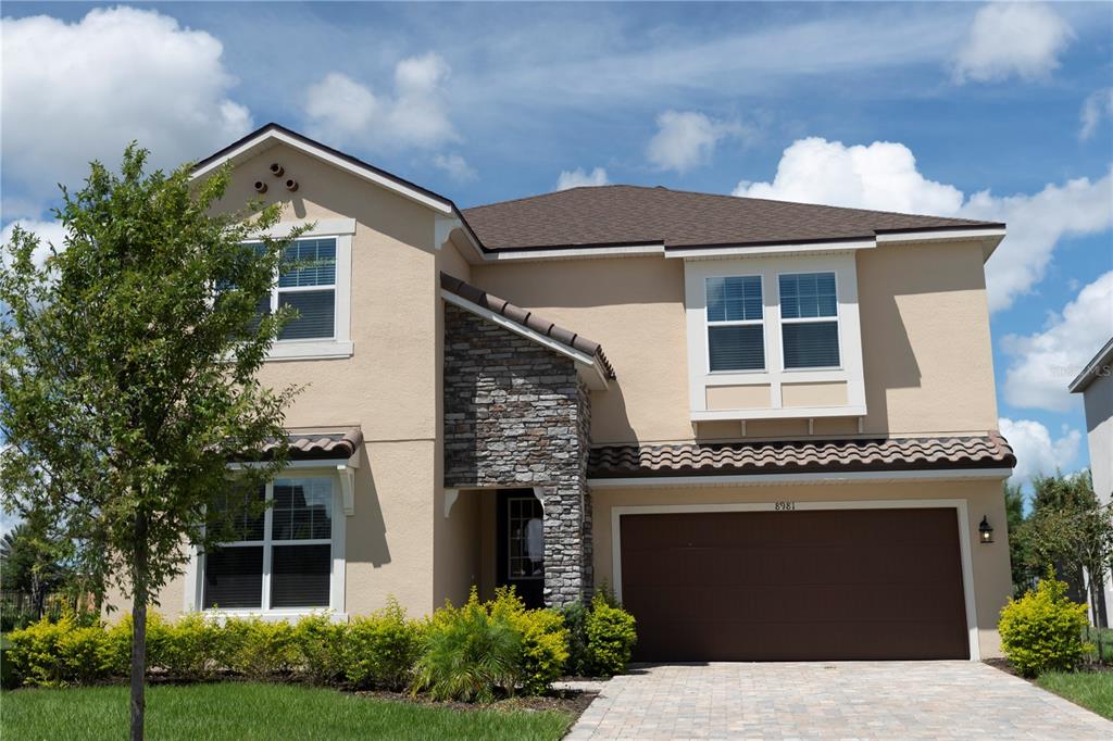 A two-story house with tan stucco, stone accents, a brown garage door, and a shingled roof — 8981 AMELIA DOWNS TRAIL, KISSIMMEE, FL