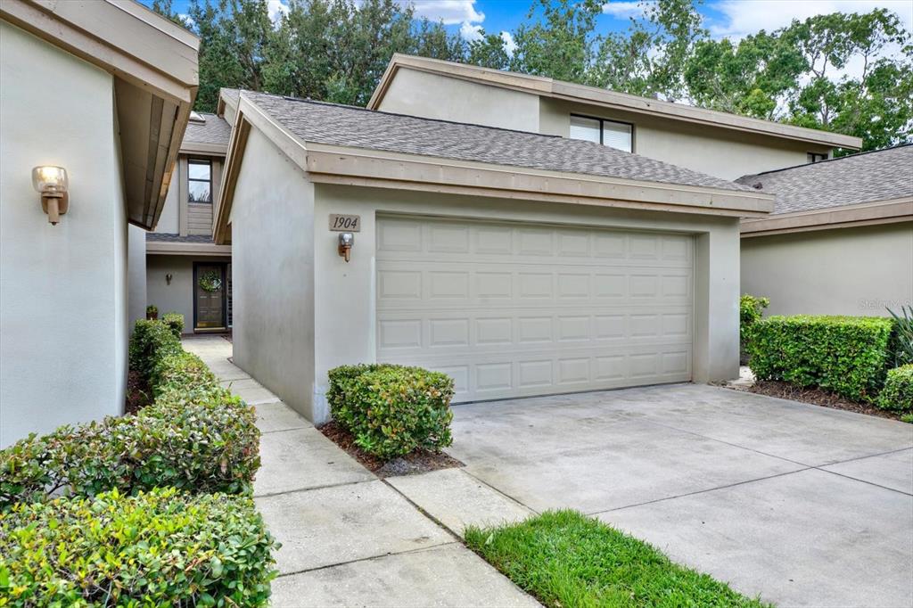 A beige house exterior features a single-car garage, a concrete driveway, and a landscaped walkway — 3090 LANDMARK BOULEVARD UNIT 1904, PALM HARBOR, FL