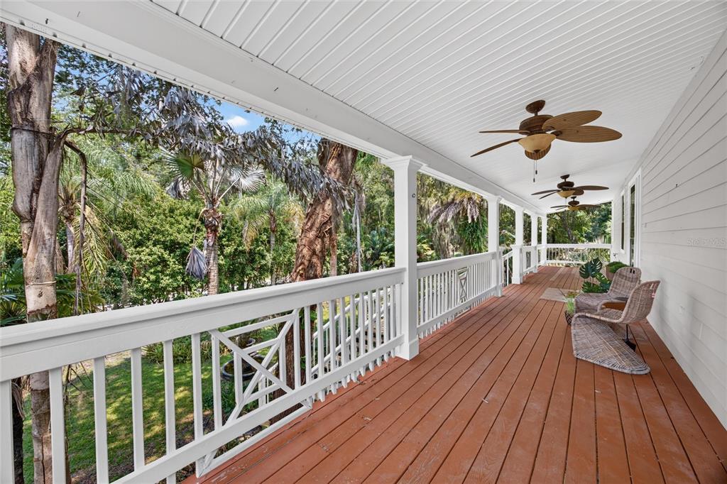 A long, covered wooden porch features white railings, ceiling fans, and wicker chairs overlooking trees — 8435 FLORENCE COVE ROAD, ST AUGUSTINE, FL