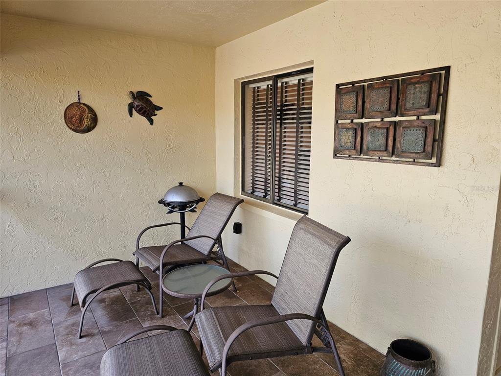 A covered patio features tiled flooring, two chairs with ottomans, a small table, and wall decor — 6468 SEAGULL DRIVE UNIT 316, BRADENTON, FL