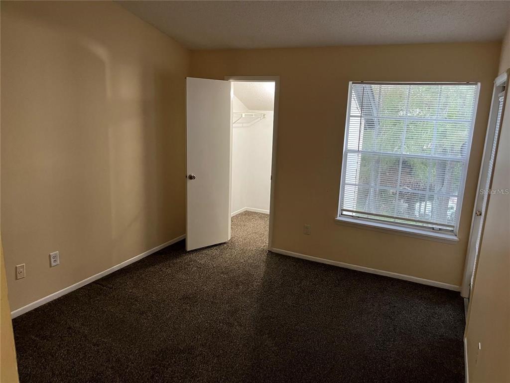 A bedroom with beige walls, dark carpet, a window with blinds, and an open closet door — 1924 LAKE ATRIUMS CIRCLE UNIT 55, ORLANDO, FL