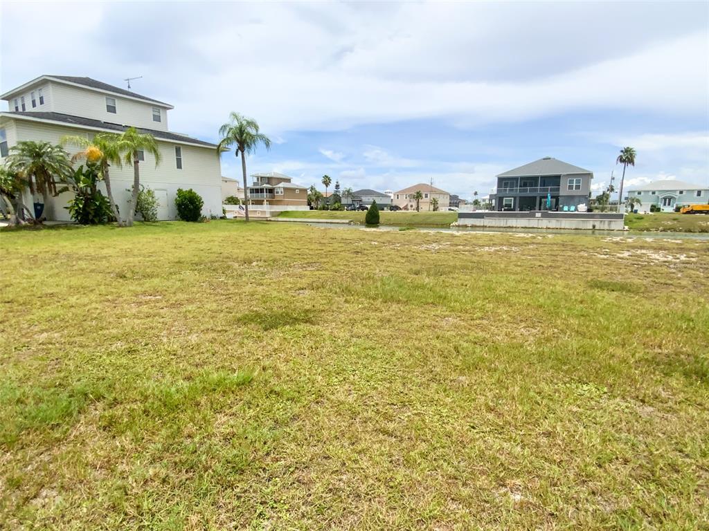 A vacant, grassy waterfront lot sits between two multi-story houses under a cloudy sky — 3471 CROAKER DRIVE, HERNANDO BEACH, FL