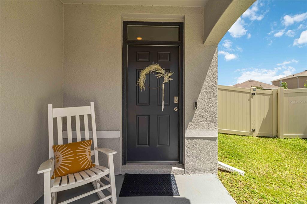 A white rocking chair sits on a concrete porch in front of a dark door — 8354 HARDY BAY LOOP, WESLEY CHAPEL, FL